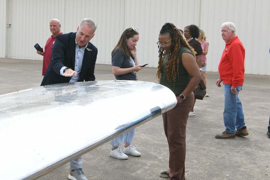 Several people gathered around a metallic airplane wing, examining it closely. A man in a suit points at something.