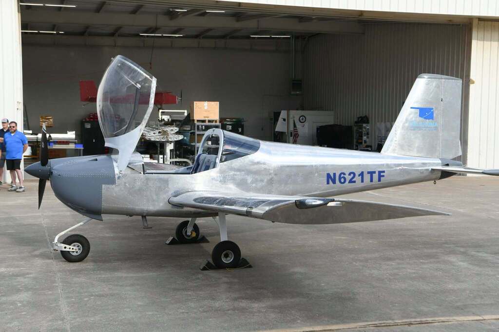 Small airplane with "N621TF" on tail, parked in hangar. Open cockpit, passengers nearby.