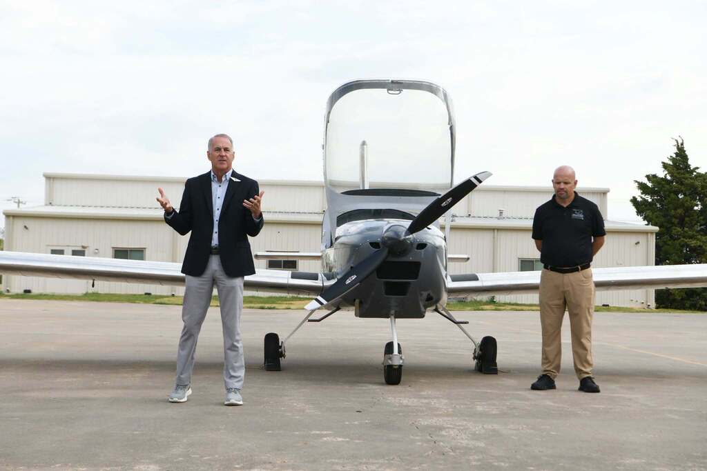 Two men stand beside a small plane on a runway. One gestures, and the other stands near the cockpit. A building with windows is behind them.