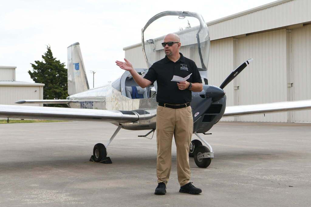 Person in sunglasses and black shirt stands in front of airplane, gesturing with hand while holding paper.