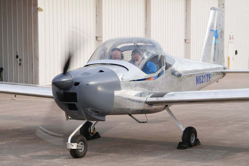 Two people sit inside a small airplane with a propeller. It stands on a runway in front of a hangar.