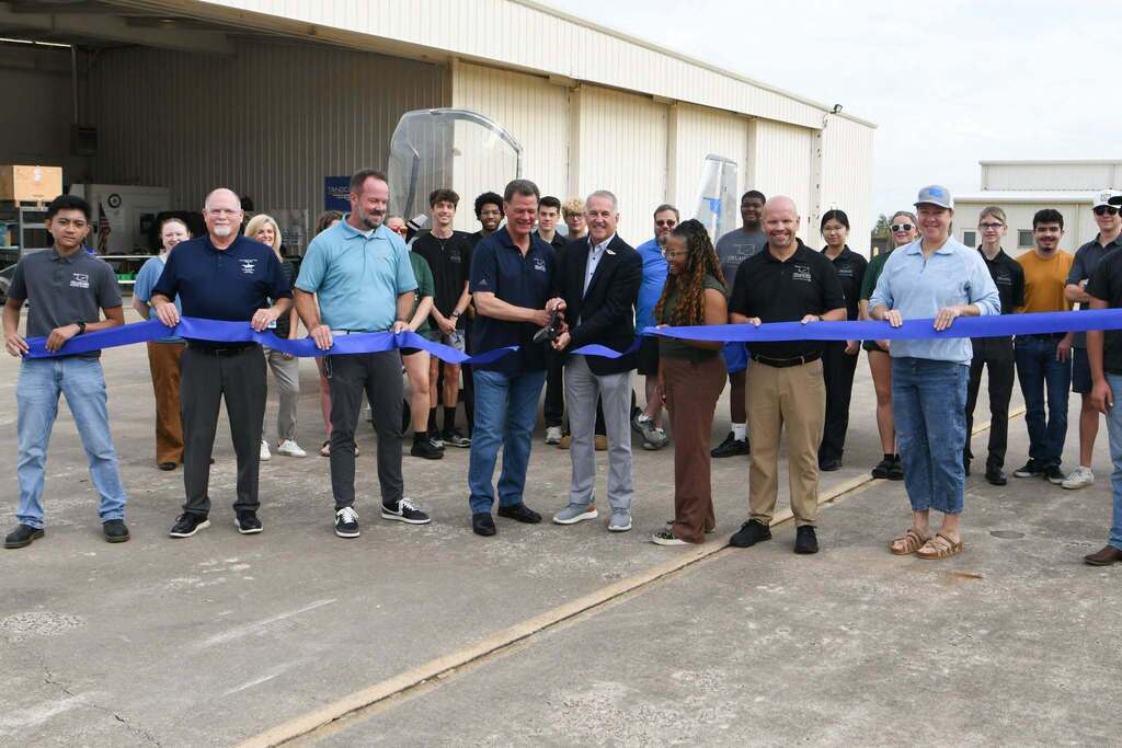Group of people standing in front of a hangar, holding a blue ribbon for an opening.