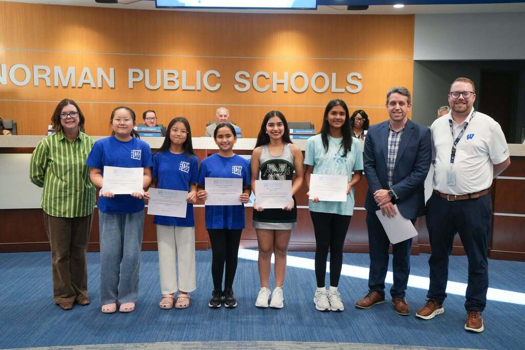 Five students holding certificates stand with three adults in a room with a Norman Public Schools sign.