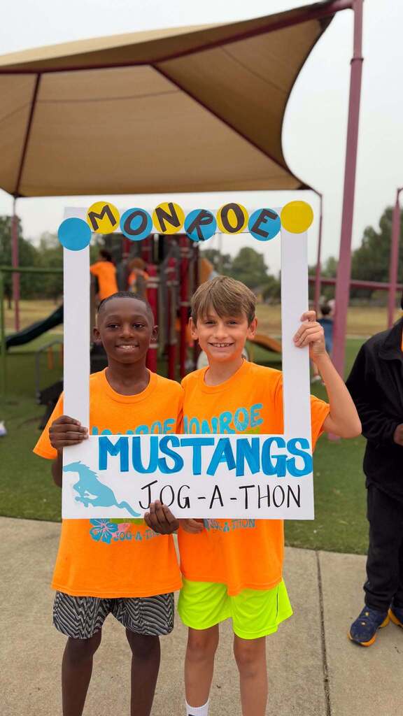 Two boys in orange shirts stand next to a photo frame reading "MONROE MUSTANGS JOG-A-THON" in front of a playground.