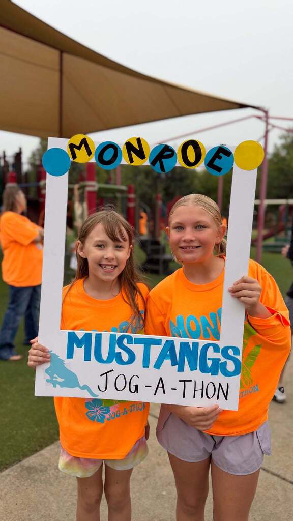 Two young girls in orange shirts hold a photo frame reading "Monroe Mustangs Jog-a-thon" in front of a playground.