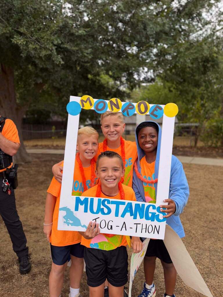 Four children pose in front of a sign, wearing orange shirts. They stand behind a sign that reads "MONROE MUSTANGS JOG-A-THON."