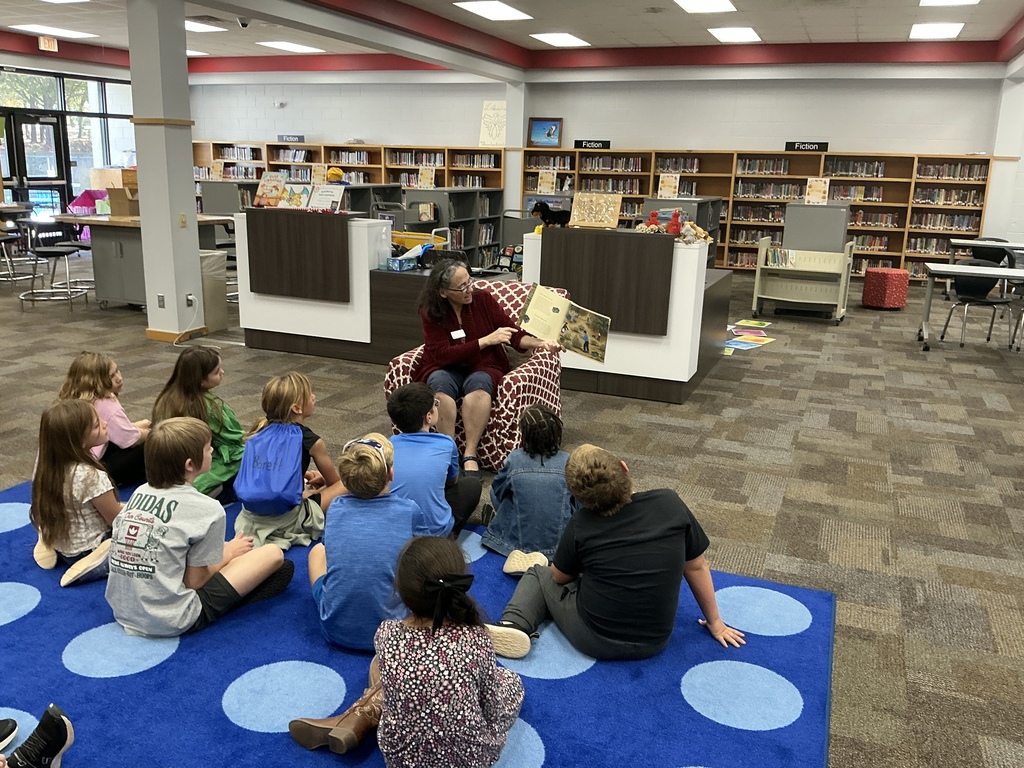 Children and an adult sit in a circle on blue carpet in a library. Behind them, bookshelves line the walls.