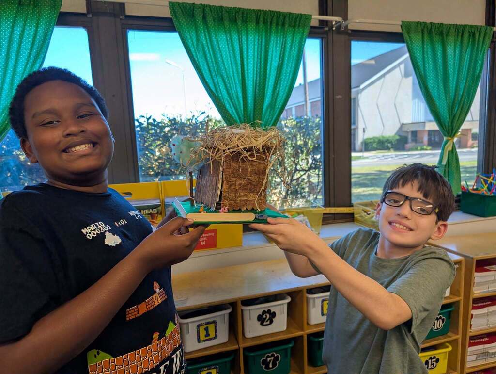 Two children in a classroom, one holding a birdhouse, both smiling. Shelves and windows with green curtains in the background.