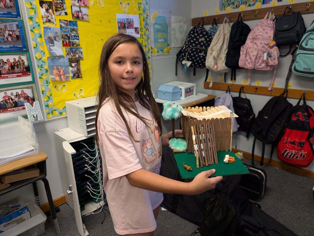 A girl holding a small architectural model in a classroom, surrounded by shelves with backpacks.