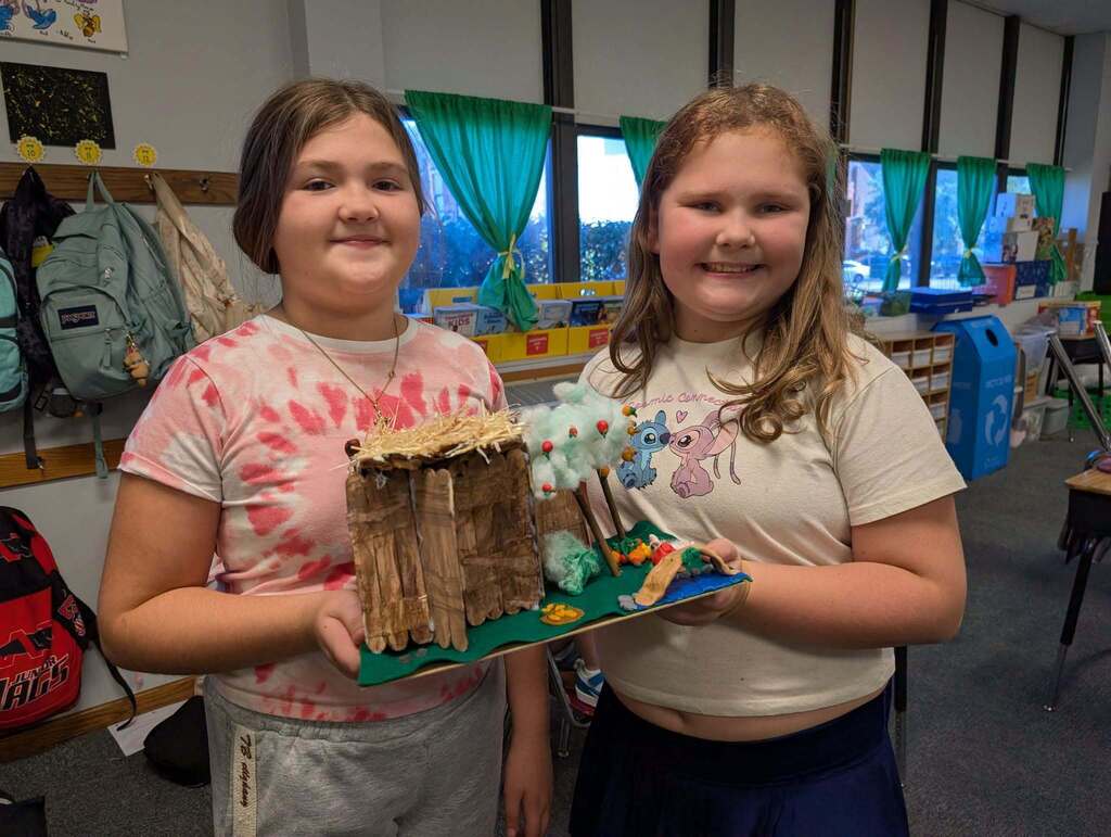 Two girls stand in a classroom, smiling, holding a handmade structure. The background includes windows and classroom items.