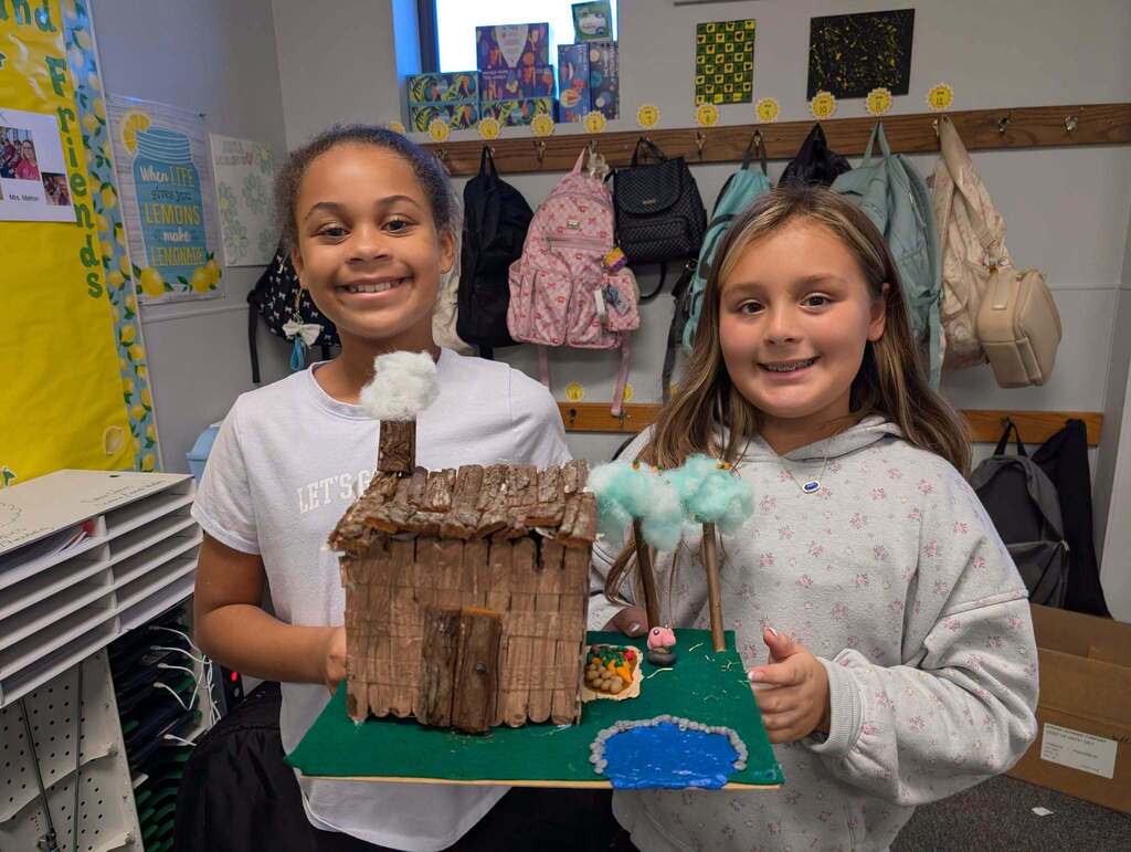 Two girls, one in a white shirt and the other in a gray hoodie, hold up a model house.