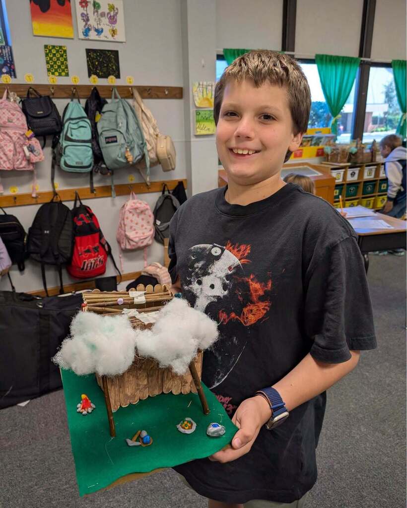 A young boy in a classroom holds a project with a cloud-topped house, smiling. Many bags hang on the wall behind him.