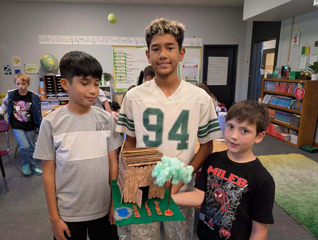 Three boys stand in a classroom with a science project. One holds a small, model volcano.