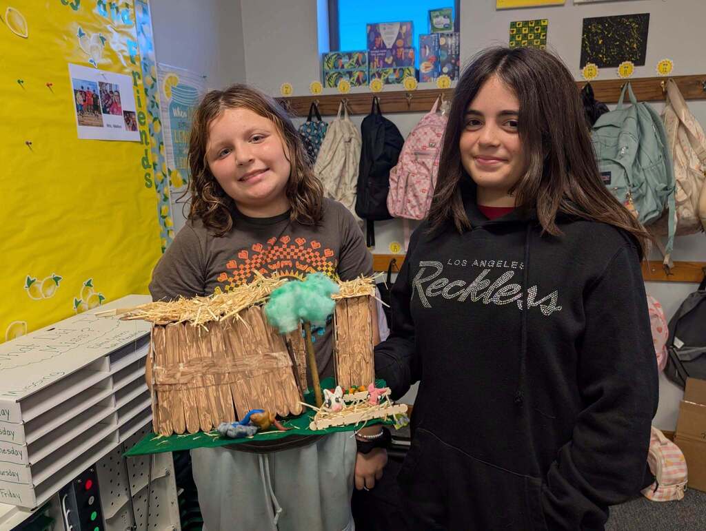 Two girls, one holding a model, pose next to a classroom wall. The model resembles a hut.