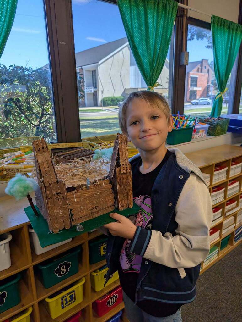 Child holding a small house-like structure made of crackers in a classroom with shelves of books.