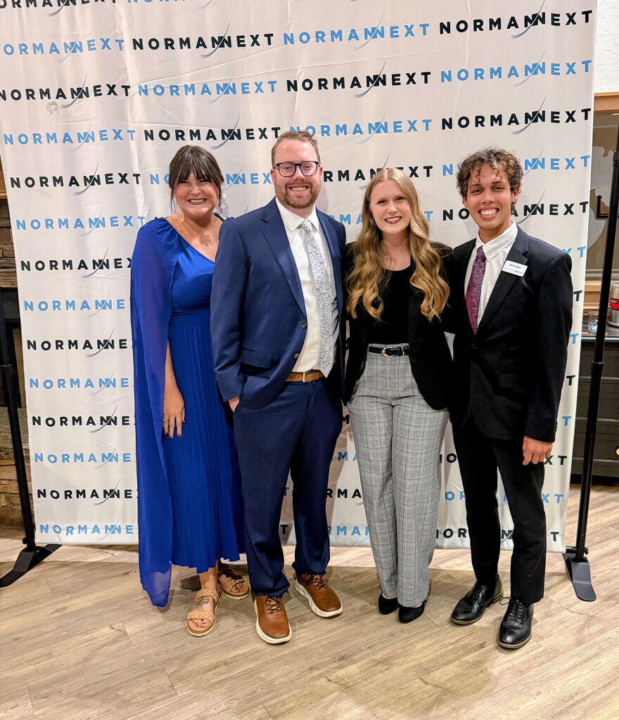 Four people stand close together for a picture. They wear business suits and appear to be smiling.