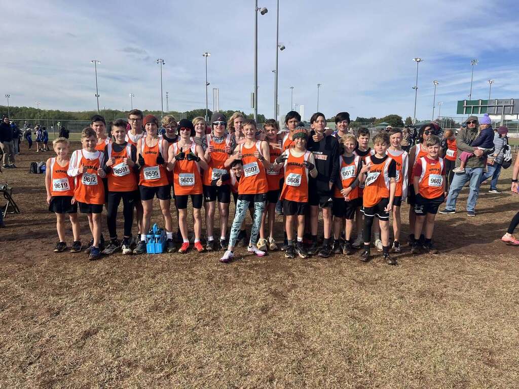 Young students in orange vests and medals stand together on a dirt field, smiling for a photo.