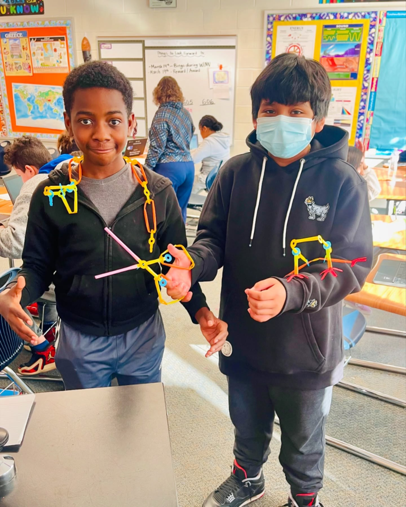Two students displaying colorful geometric structures they built using connecting pieces in a classroom.
