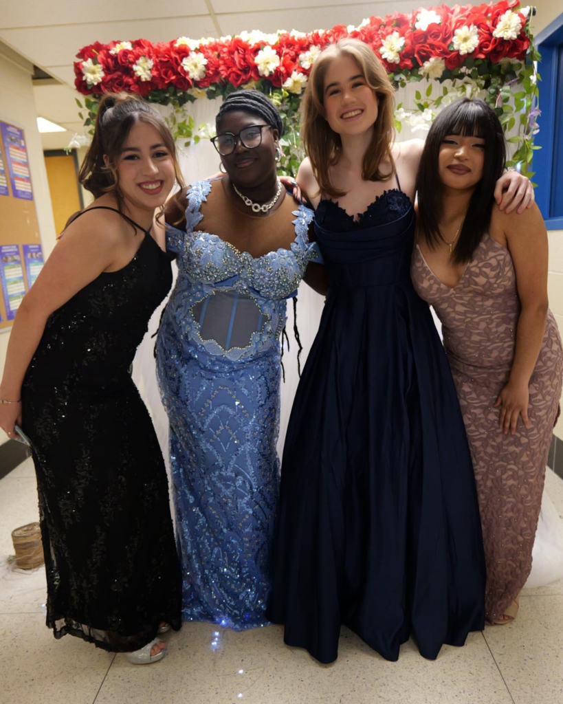 Four students at the junior prom posing for photo in front of white backdrop decorated with flowers