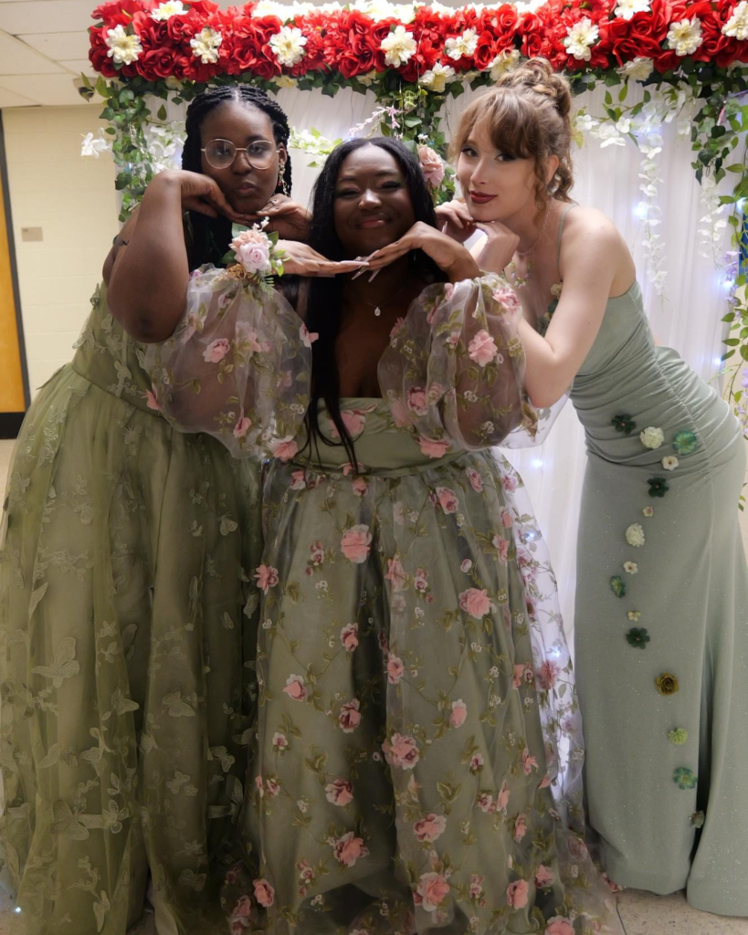 Three students at the junior prom posing for photo in front of white backdrop decorated with flowers