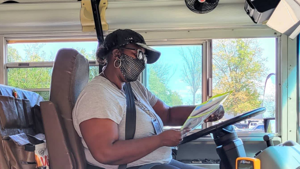 A bus driver wearing a mask sitting in the driver's seat of a school bus, reviewing papers while parked.