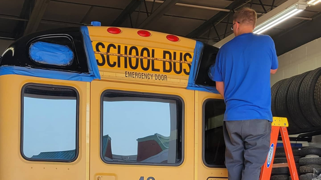 A staff member in a blue shirt working on the roof of a yellow school bus in a garage.