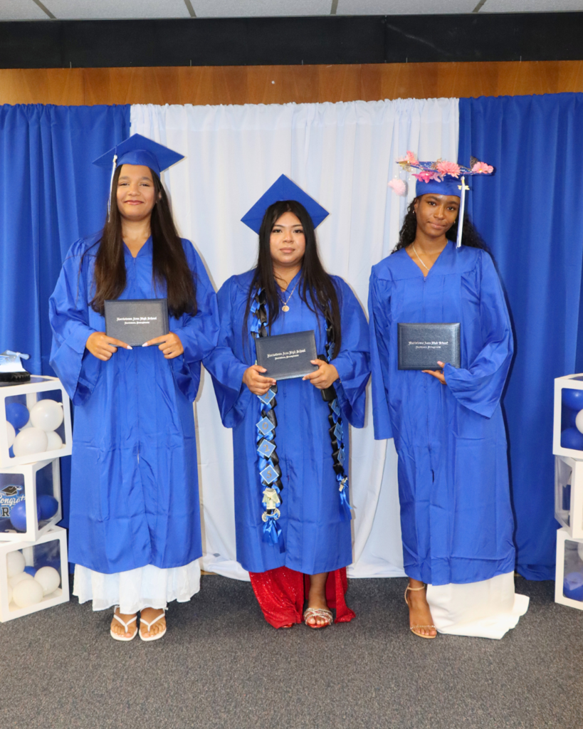 Three graduates in blue caps and gowns holding diplomas, standing in front of blue and white backdrop