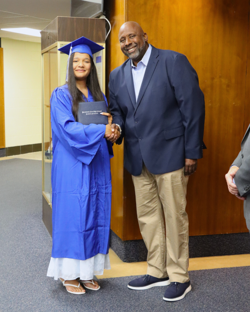 Graduate in blue cap and gown shaking hands with Dr. McGriff, principal while holding diploma
