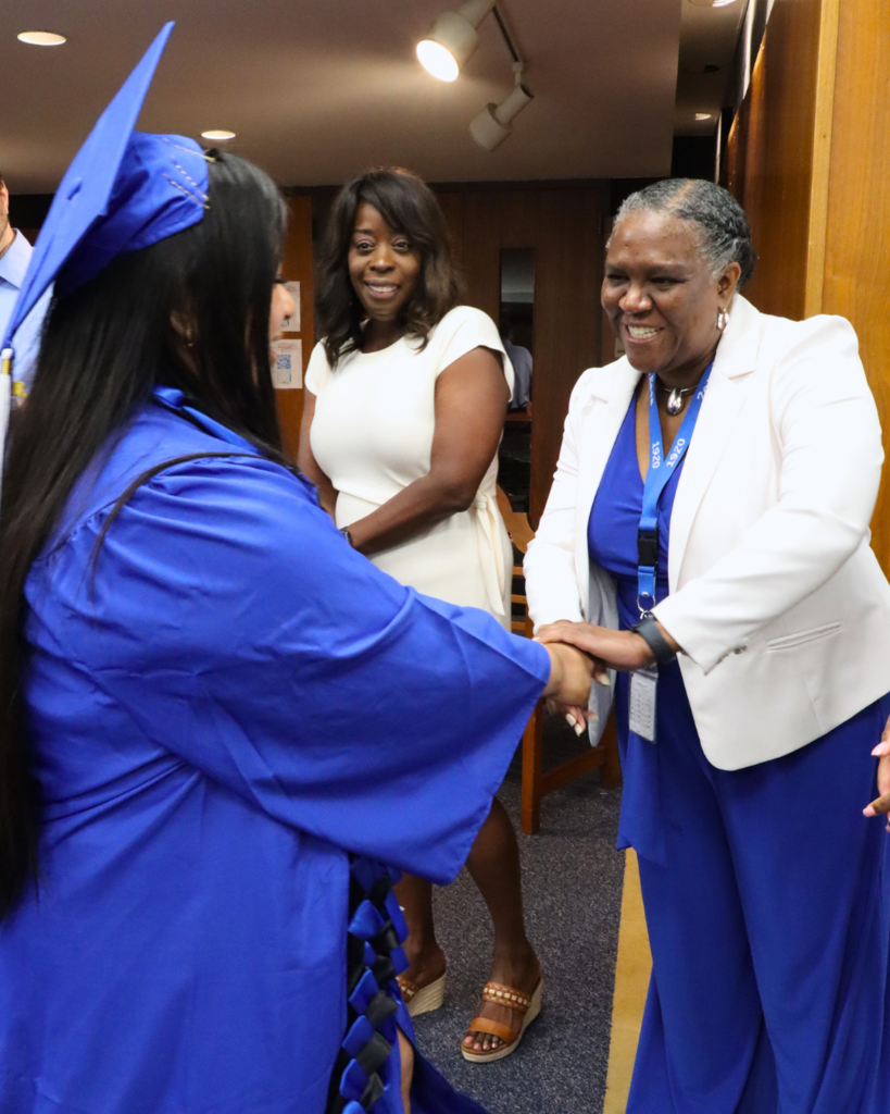 Graduate in blue cap and gown from behind, shaking hands Ms. Casimir, Assistant Principal