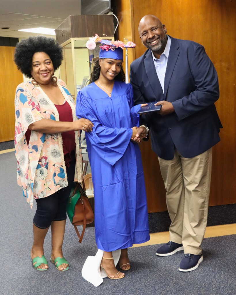 Graduate in blue cap and gown with decorative cap standing between a family member and Dr. McGriff, principal.