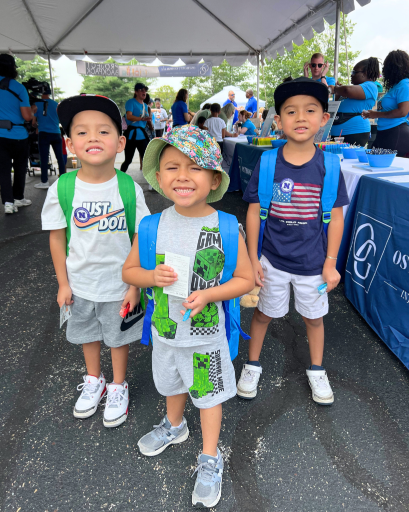 Three young students pose for photo with their free backpacks in front of the giveaway tables with volunteers in the background.