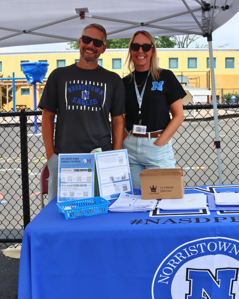 Two NASD staff members pose for photo behind NASD information table and underneath a white tent. .