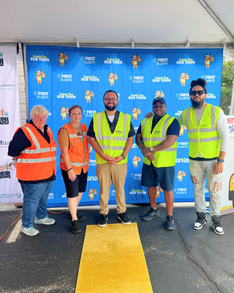 Five First Student staff members pose for photo in front of backdrop with a yellow carpet.