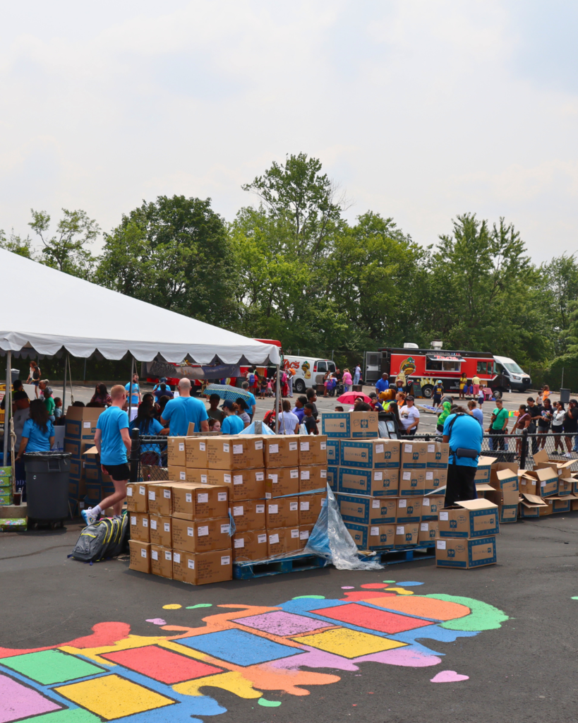 Two wooden pallets of boxes filled with backpacks and school supplies for families. Volunteers and families in the background.