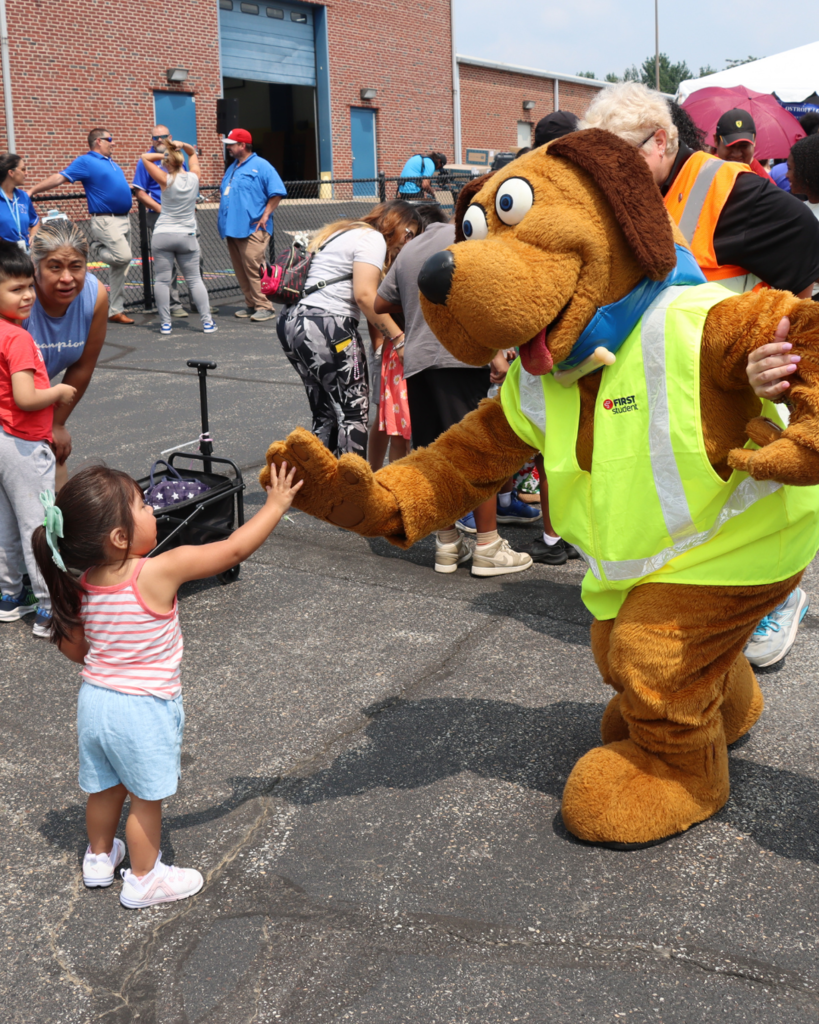 First Student Mascot, Safety Dog, leans over to give a high-five to a young girl.