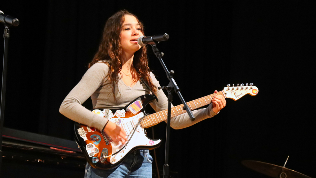 Student performer playing guitar and sining into microphone on stage.