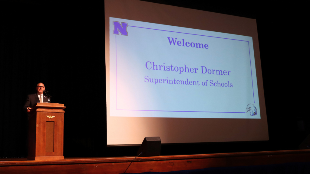 Mr. Dormer speaking at the podium on stage with "welcome" slide on screen.