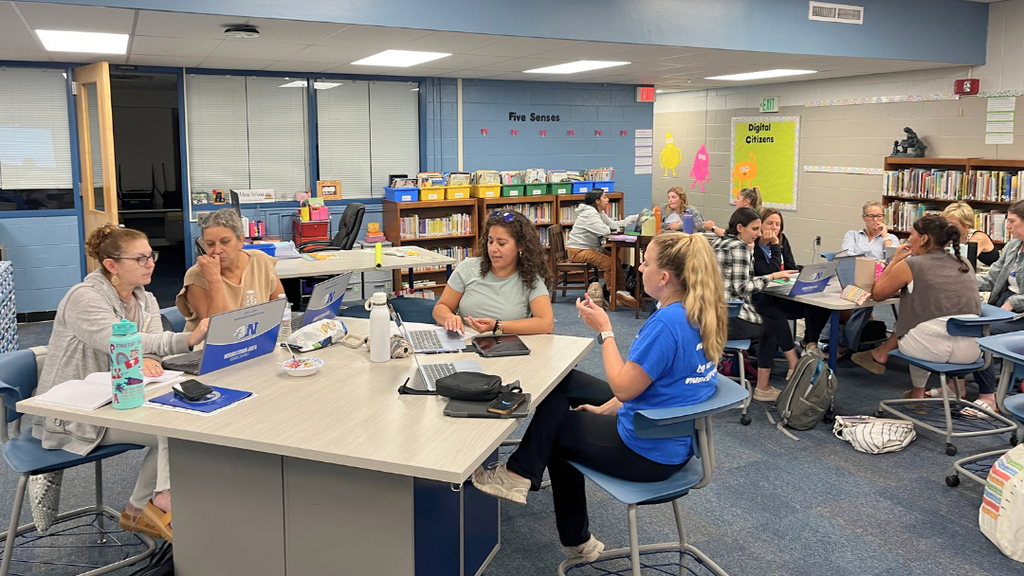 Groups of teachers sit together at tables engaging in discussion. 