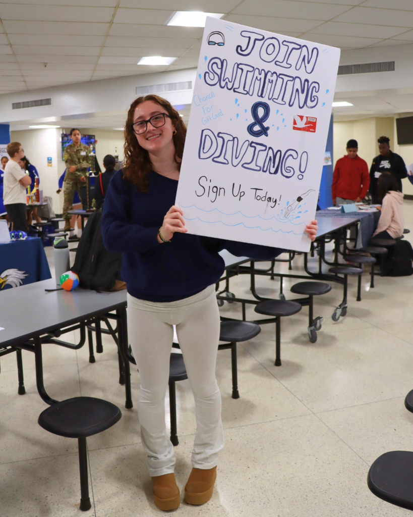 Student holding up a poster that says "Join Swimming and Diving. Sign Up Today" in the cafeteria.