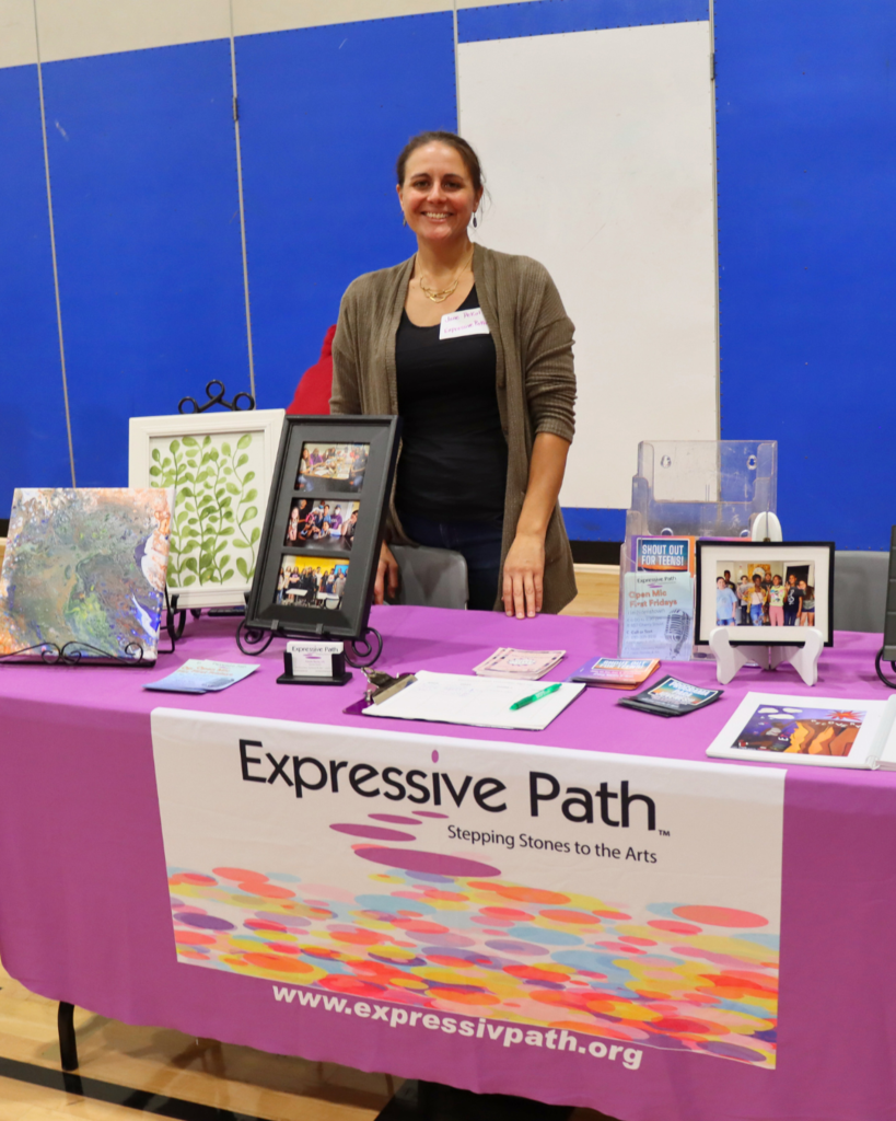 Community partner poses for photo behind their table with photos and items on display.