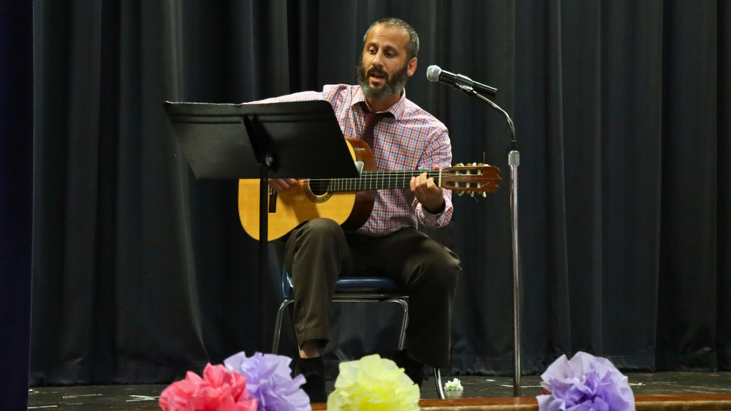 Man in a chair playing guitar on stage with a music stand and microphone.