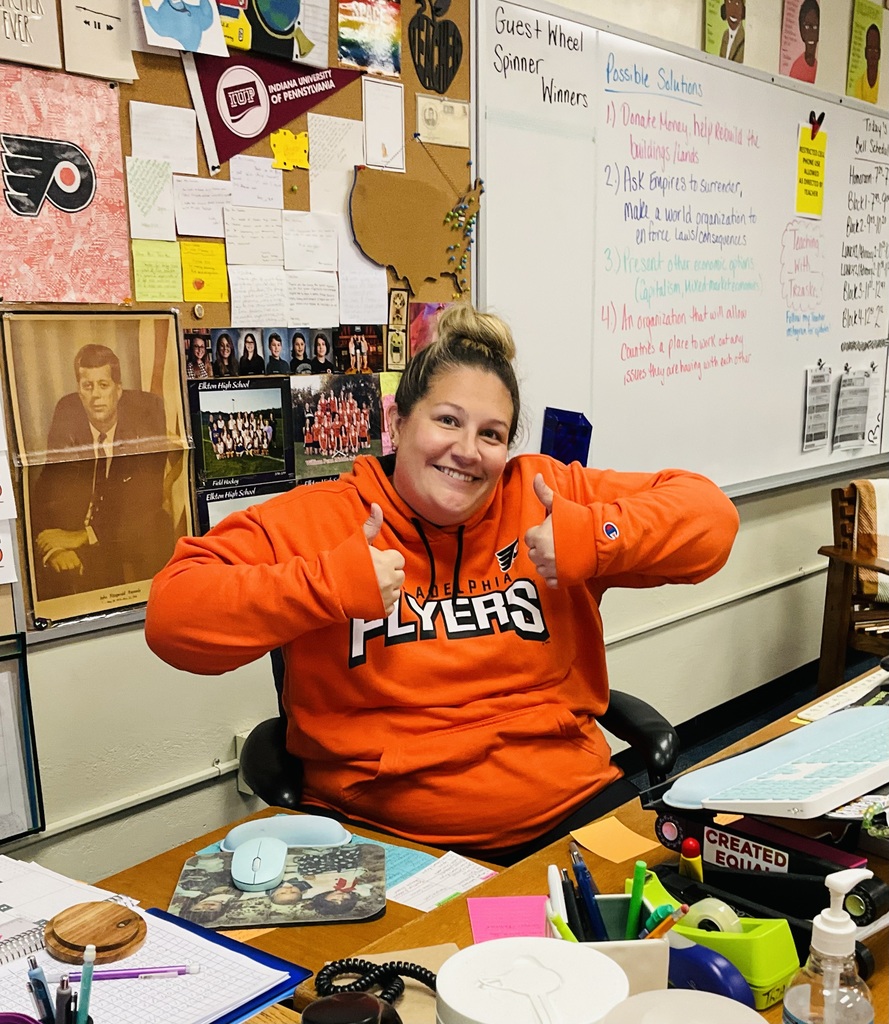 A teacher is seated at her desk in her orange clothes and she is giving a thumbs up