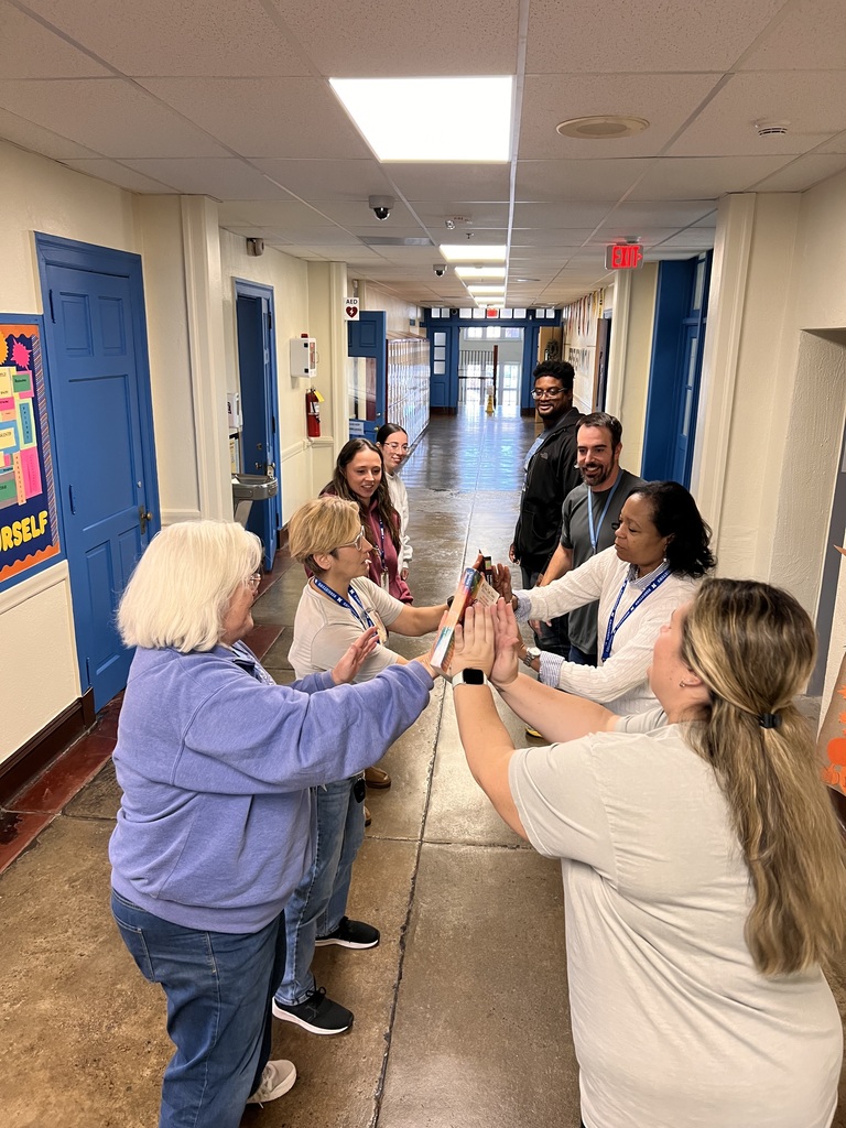 Eight teachers are doing a brain break in the hallway with passing a book with their just their palms