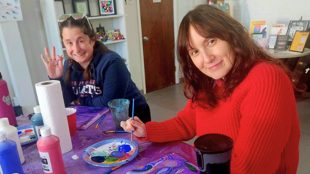 Two teachers working on their paintings smile for a photo.