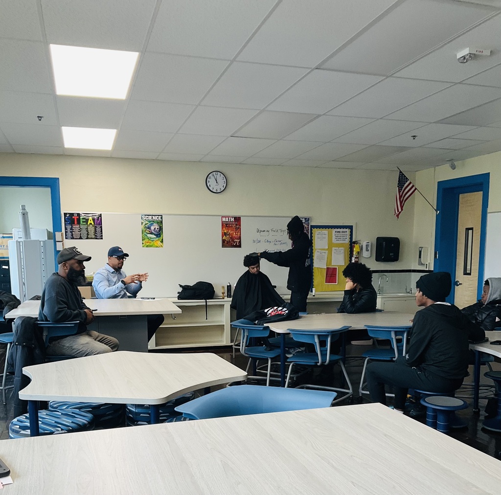 A photo of a student getting his hair cut while other students are listening to the presentation
