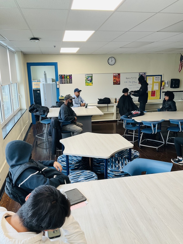 A photo in the classroom of students listening to the presenters talk while a student is getting his hair cut by another barber.
