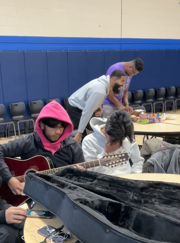 A student is playing the guitar with teachers laughing in the background as they are playing games