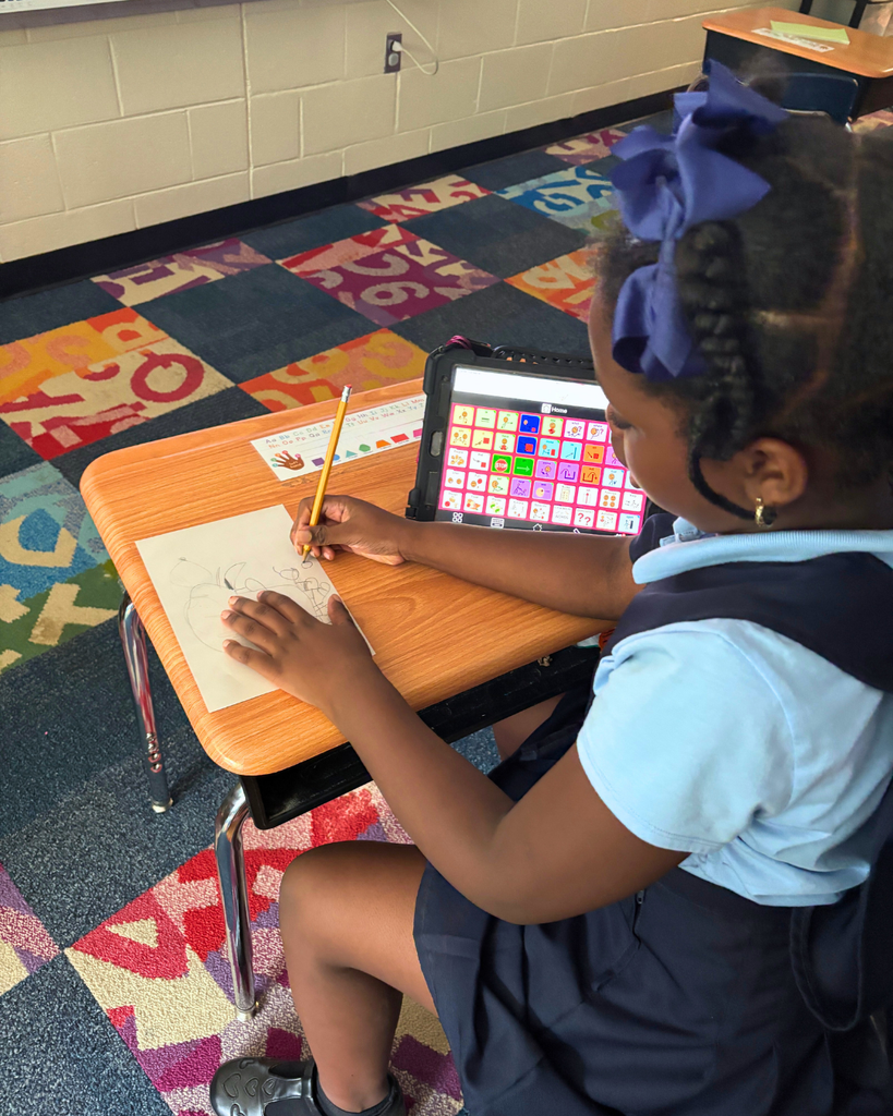 Student drawing at desk with their Augmentative and Alternative Communication (AAC) device. 