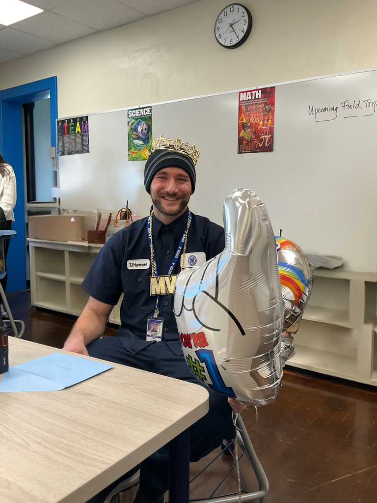 The custodian is posing in a classroom with balloons and a crown
