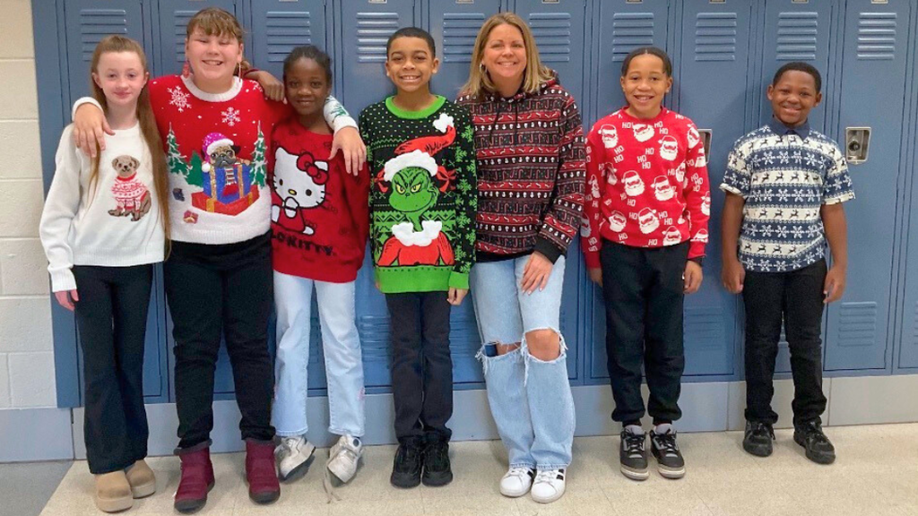 Teacher and students wearing fun holiday sweaters pose for a photo in front of lockers. 